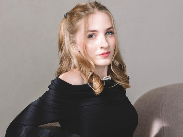 Eva Daly, con coletas rubias, ojos azules y labios rosados, vestida con una blusa negra con un hombro descubierto, mostrando sus pequeños senos y su linda sonrisa, sentada en una silla gris..