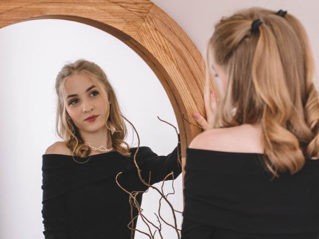 EvaDaly with wavy blonde hair in a black off-shoulder top, looking into a wooden mirror, showing her reflection with a slight smile and a pearl necklace.