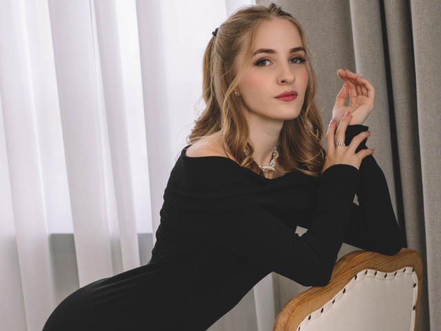 EvaDaly with blonde hair, black off-shoulder dress, and red lipstick, leaning on a chair, hand raised, looking at camera, wearing a necklace, soft light, elegant.