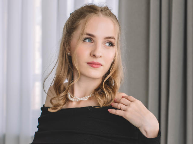 EvaDaly with blonde hair, blue eyes, and pink lips, wearing a black off-shoulder top and pearl necklace, slightly pulling top with right hand, looking to the side.