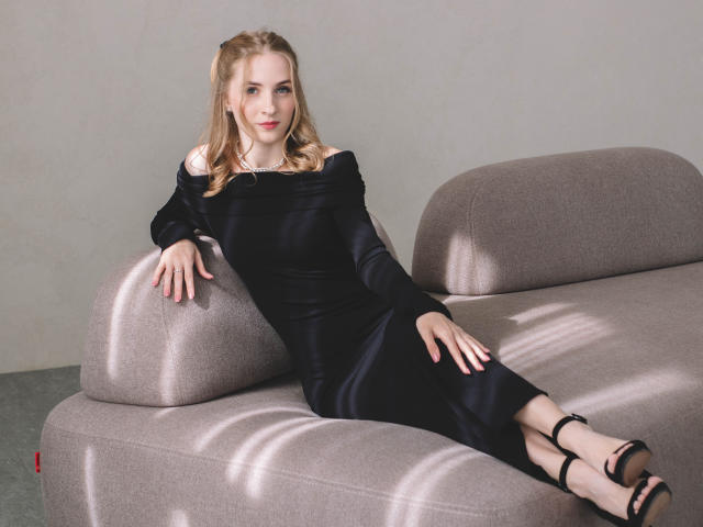 EvaDaly with blonde hair, wearing a black off-shoulder dress and black strappy heels, lounging on a gray couch, showing her tits and ass, with a confident smile.