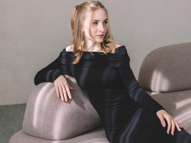 EvaDaly with blonde pigtails, wearing a black off-shoulder dress, sitting on a beige sofa, her hand on the armrest, her other hand on her knee, looking sideways.