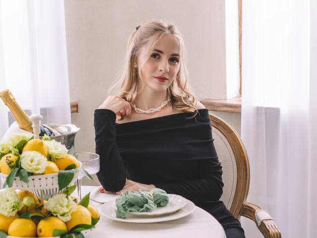 EvaDaly with blonde hair, off-shoulder black dress, pearl necklace, sitting at table, lemon bouquet, champagne, green napkin, wooden chair, window with white curtains, soft smile.