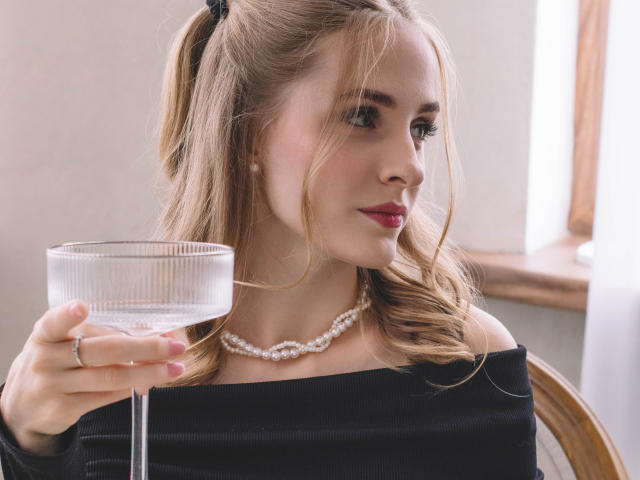 EvaDaly with wavy blonde hair in a black off-shoulder dress, holding a glass of white wine, wearing a pearl necklace, and red lipstick, looking to the side.