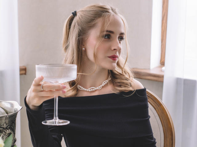EvaDaly with blonde pigtails, off-shoulder black dress, pearl necklace, holding a clear glass of wine, sitting, looking to the side, red lipstick, wooden chair, soft light.