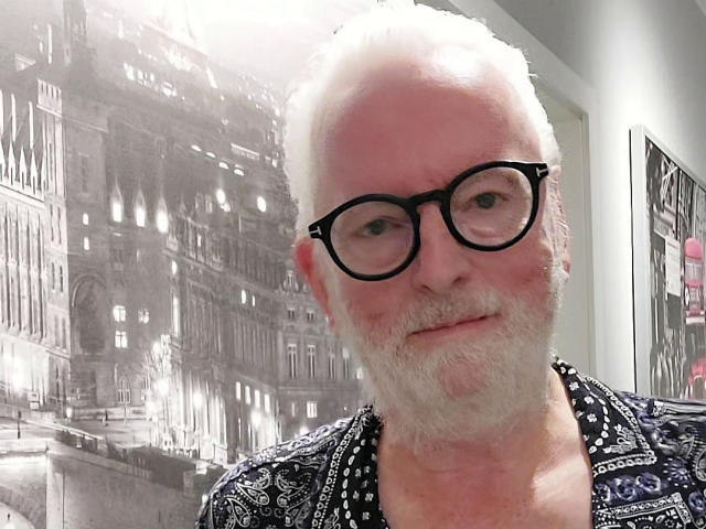 JackGreyHot é um homem mais velho, com cabelo branco, óculos e barba, vestindo uma camisa estampada, sorrindo levemente e com o rosto um pouco avermelhado..