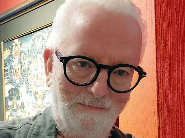 JackGreyHot ha i capelli bianchi, una barba, occhiali neri e un leggero sorriso. Indossa una camicia grigia ed è in piedi di fronte a un'opera d'arte colorata, incorniciata..