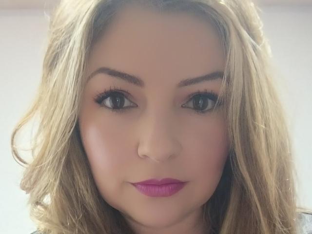 NatalyHami's face with blonde wavy hair, pink lipstick, and thick eyebrows, showing her green eyes and subtle makeup. Her nose is small and her skin is light.