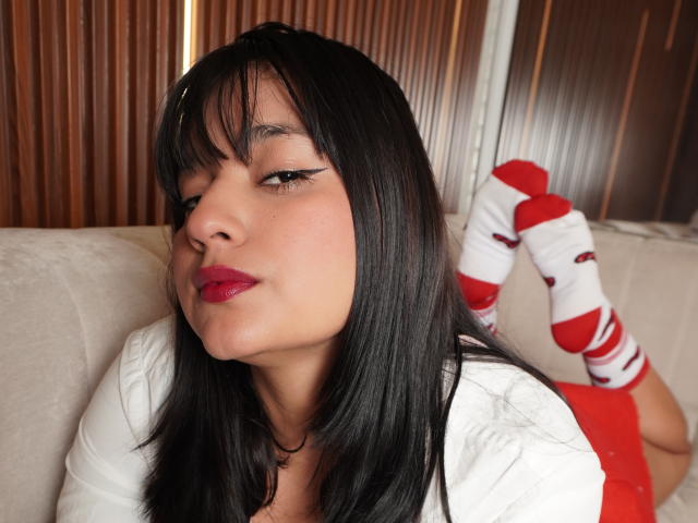 MiaEverly has black straight hair, red lipstick, and is lying on a couch with white socks and red stripes, wearing a white top, showing her ass.