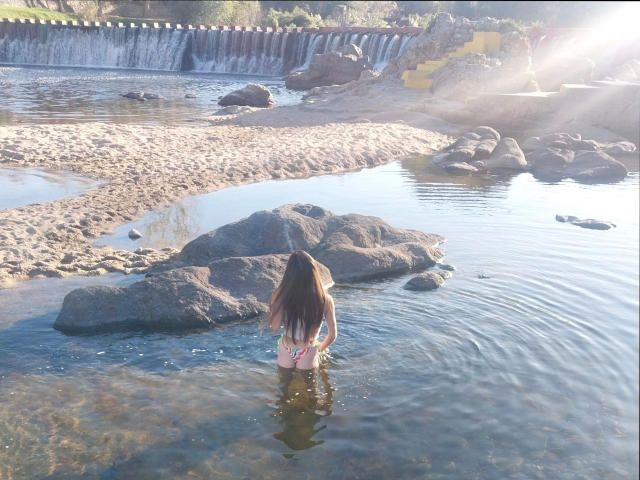 GimenaCat-hot stands in shallow water with long brown hair, wearing a bikini, facing rocks. Sunlight flares from the upper right. GimenaCat-hot's back is to the camera.