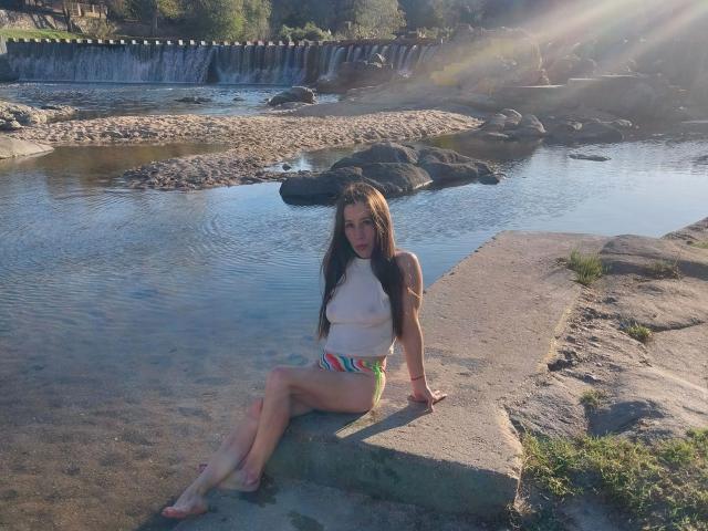 GimenaCat-hot with long brown hair, wearing a white top and colorful bikini bottoms, sits on a stone ledge by a shallow, rocky stream, sunlight shining through.