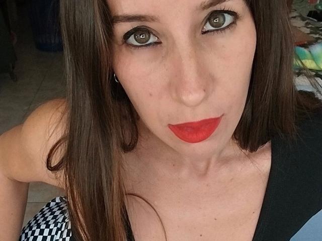 GimenaCat-hot has green eyes, red lipstick, and brown hair, with a black and white patterned top, looking up with a slight pout. Her tits are partially visible.