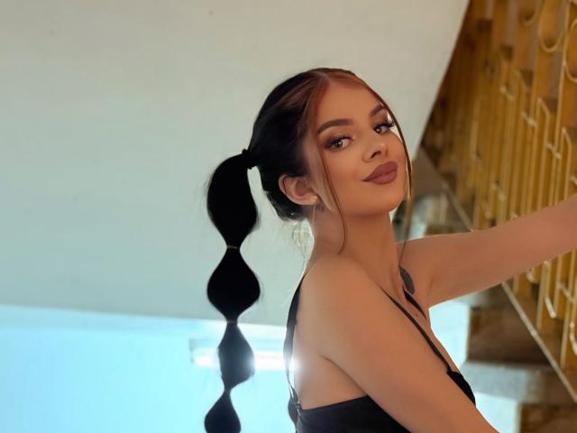 IvyRaven with pigtails, black bra, and lipstick, smiling, showing her tits, standing next to a golden staircase, under soft light.