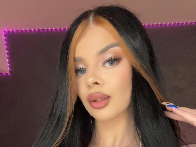 IvyRaven has long black hair with blonde highlights, blue eyes, and pink lips. She's holding her hair, showing blue nails. Purple LED strip lights in background.