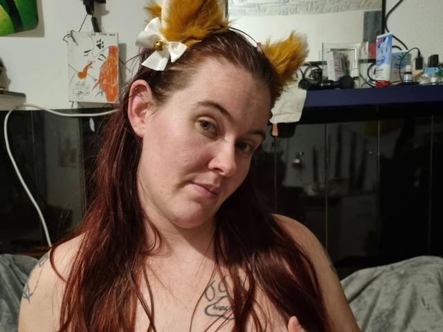 Rotkaeppchen-hot with brown hair, yellow furry cat ears, white bow, and tattoos, looking at camera with a slight smile, topless, showing her bare tits, in a dimly lit room.