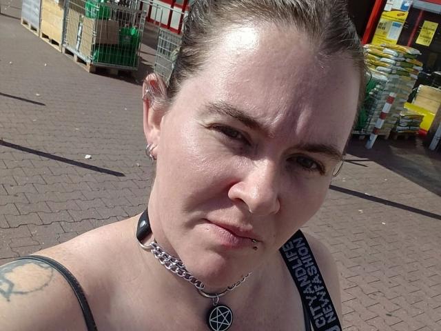 Rotkaeppchen-hot with a pensive look, light skin, brown hair in a bun, small nose ring, black choker with pentagram, tattoo on shoulder, standing outside in a sunny parking lot.