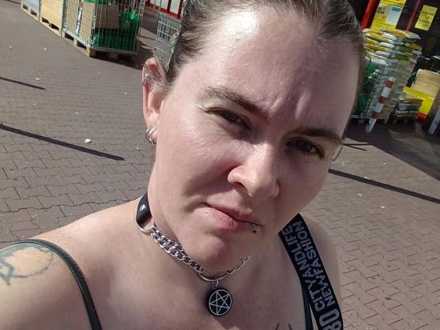 Rotkaeppchen-hot with a nose ring, lip piercing, and pentagram necklace, wearing a black strap top, has a tattoo on her left shoulder and a chain collar.