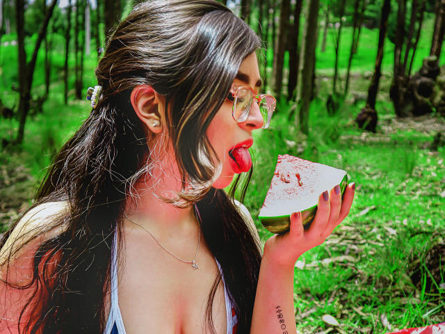 EvaMcAllister with long dark hair, glasses, and red lipstick, sticking out her tongue while eating a slice of watermelon, showing her small tits and tattoo on her right wrist.