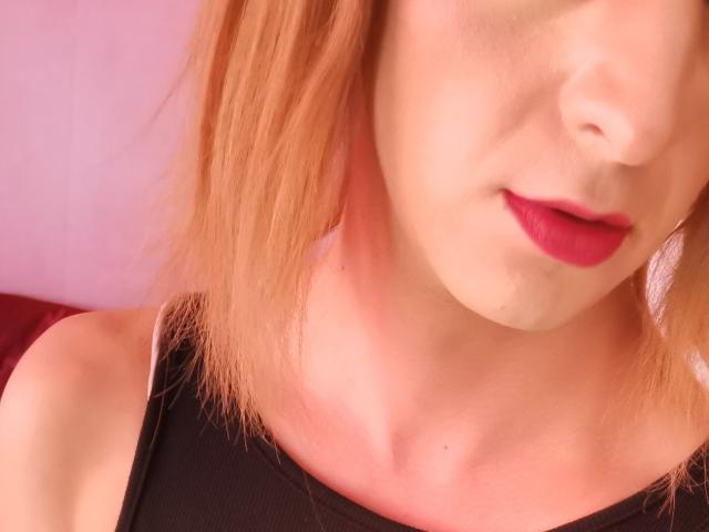 MiaMystique's face, showing a slight smile with red lipstick, bare shoulders, and a black top, with her blonde hair and fair skin in a soft pink light.