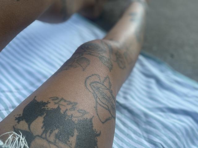 DaneeSoHot's tattooed leg with black ink tattoos on dark brown skin, lying on a striped towel, showing a flower and wolf tattoo on the upper thigh.