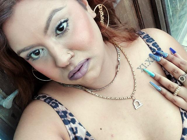 GoddessLorenaTheeSiren with pinkish skin, blue-green eyes, purple lipstick, gold hoop earrings, chain necklace, leopard print top, blue and green nails, heart tattoo, and ring, looking at camera.