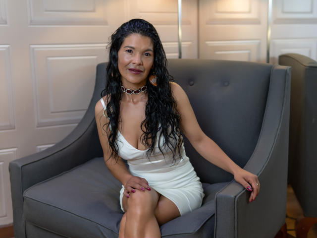 MollyOrtega with long black wavy hair, wearing a white dress, sitting on a gray armchair, smiling, showing her tits, and wearing a black choker with red gemstones.