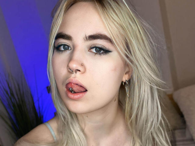 IriskaMimi with blonde hair, blue eyes, black eyeliner, nose ring, tongue piercing, and gold hoop earrings, sticking out her tongue. Her skin is fair with a small blemish.