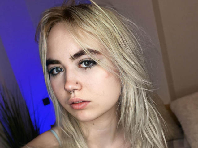 IriskaMimi has blonde hair, blue eyes, and a nose ring. She's looking at the camera with a slightly pouty expression, wearing a light blue top.