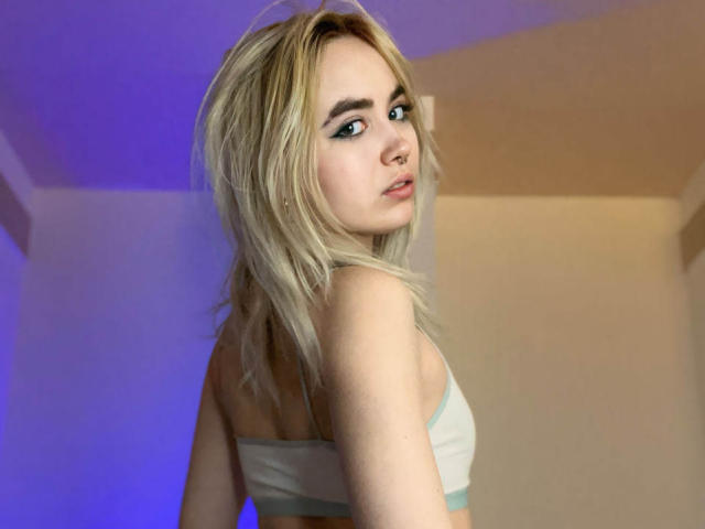 IriskaMimi with blonde hair, blue eyes, nose ring, and white bra, looking over shoulder, showing her tits and ass in a slightly blurred, dimly lit photo.