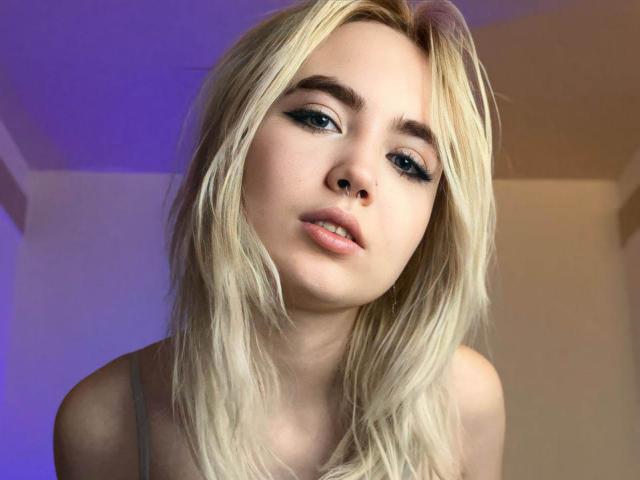 IriskaMimi with blonde hair, blue eyeliner, and parted lips, showing a slight pout. She's wearing a grey top, and her face is centered in the frame.