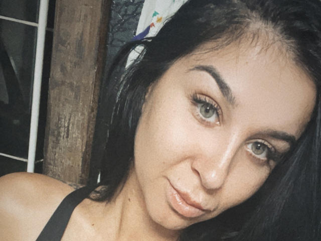FrancescaDante has dark black hair, light brown skin, green eyes, and is pouting with her glossy lips. She's wearing a black top, showing her bare shoulders.