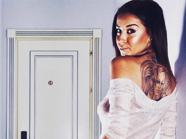 FrancescaDante with dark hair, looking back, showing large tattoo on right shoulder, wearing off-shoulder white top, in bright room, white door in background.