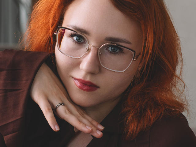 TeresaBirt with red hair, glasses, and red lipstick, wearing a brown suit, has her hand near her chin, showing a ring on her finger. Her blue eyes are focused.