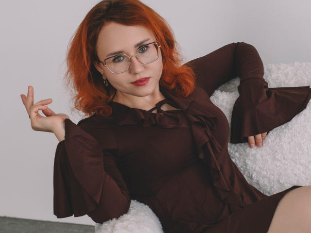 TeresaBirt with red hair, glasses, and red lipstick, wearing a brown dress with ruffled sleeves, sitting on a white fluffy chair, showing off her tits and confident smile.