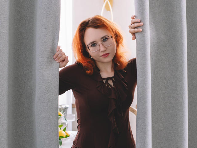 TeresaBirt with red hair, glasses, and a brown dress, holding gray curtains, standing between them, looking slightly to the side, with a subtle smile, medium-sized tits.