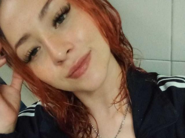 SamanthaCollen with red wet hair, blue eyes, and long lashes, wearing a navy track jacket with white stripes, smiling softly, her hand touching her head.