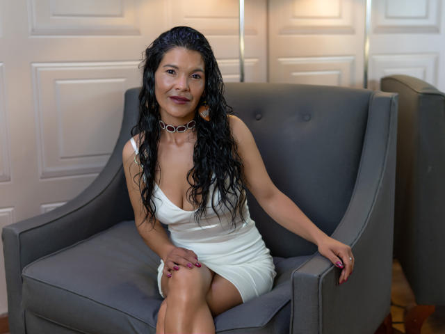Molly Ortega, con el cabello negro y mojado, vestida con un vestido blanco, sentada en un sillón gris, con uñas moradas, un collar, sonriendo, con las manos sobre el sillón, mostrando su trasero, en una habitación acogedora..