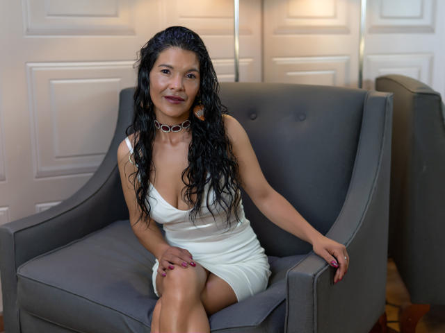 Molly Ortega, con cabello negro largo y ondulado, vestida con un vestido blanco ajustado, sentada en un sillón gris, mostrando sus senos, sonriendo, con un collar, en una habitación acogedora..