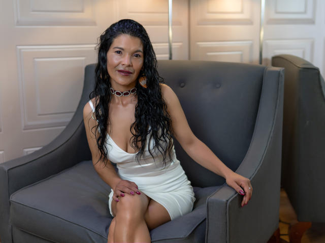 Molly Ortega est assise dans un fauteuil gris, vêtue d'une robe blanche à décolleté plongeant qui révèle ses seins, avec de longs cheveux noirs bouclés, un collier et des ongles rouges..