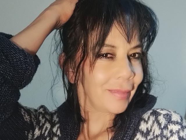 SamaraBellet with messy black hair, holding it up, wearing a knitted, dark-and-white-patterned sweater, smiling subtly, brown eyes, fair skin, and a slightly flushed face.