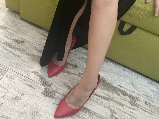 Millinia's legs in red pointed-toe heels with a black skirt, her bare thighs, and a green couch in the background on a wooden floor. Her feet are pointed.