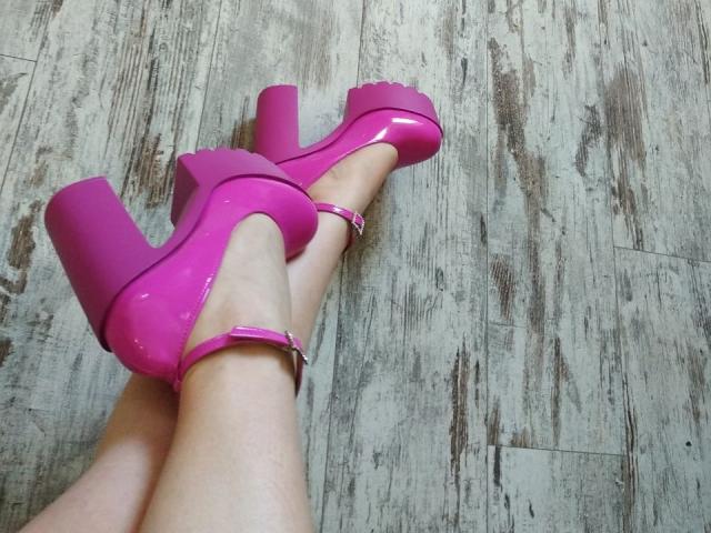 Les élégantes talons roses de Nurri, avec leurs semelles épaisses et leurs brides à la cheville, reposent sur sa peau pâle, et on voit ses jambes croisées sur un plancher en bois texturé..