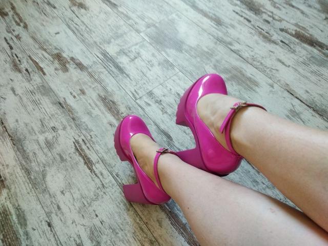 Les jambes de Nurri, chaussées de talons aiguilles roses brillants avec brides aux chevilles, sont posées sur un plancher en bois. Sa peau pâle contraste avec le rose vif de ses chaussures..