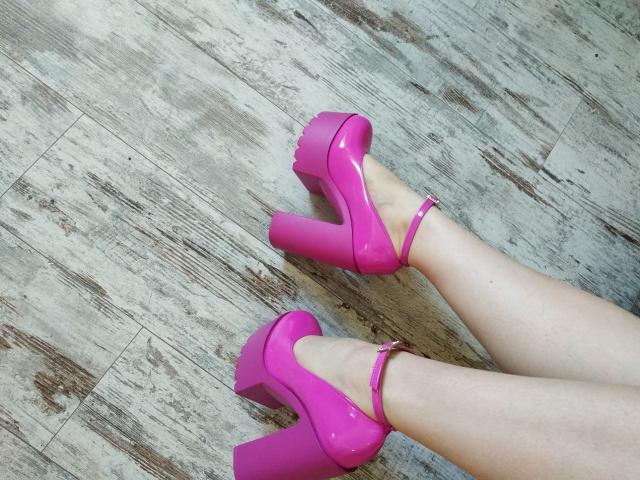Nurri's glanzende, roze hoge hakken met dikke platformsolen en enkelbandjes, staan op een houten vloer en laten haar bleke benen en roze gelakte teennagels zien..