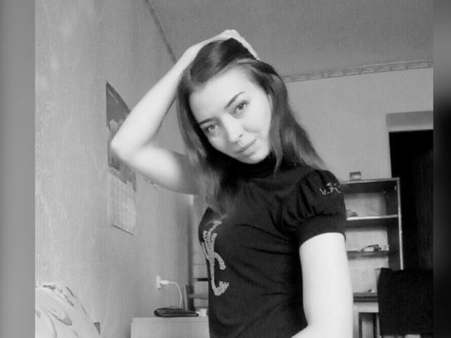 Black-and-white photo of VikkiKsu with medium-length dark hair, hand in hair, wearing a black t-shirt with a sparkly design, looking at camera with a slight smile.