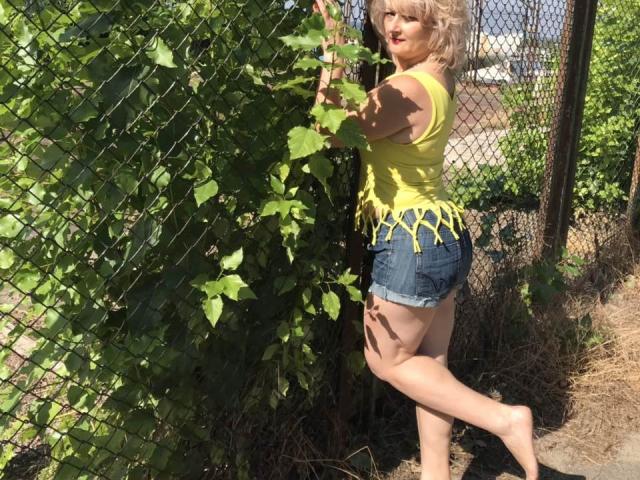 NikolinaBounty, blonde, yellow tank top with frayed hem, denim shorts, barefoot, leaning against a chain-link fence with green leaves, sunlight, red lipstick, curvy, ass visible.