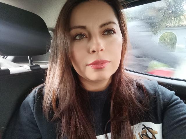 Seher has medium brown hair, light skin, and full pink lips. She's wearing a black sweater, sitting in a car with a rain-streaked window in the background.