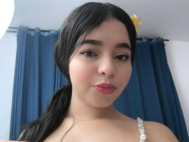 LissyCarter has medium skin, black hair in a ponytail, dark eyebrows, winged eyeliner, pink lipstick, and a yellow flower hairpin. She's wearing a bead necklace and a white top.