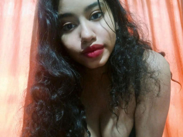 SaraMonrroe with curly black hair, red lipstick, and a slight smile, looking down, showing cleavage, against an orange curtain. Her tits are visible, and she's wearing a black top.