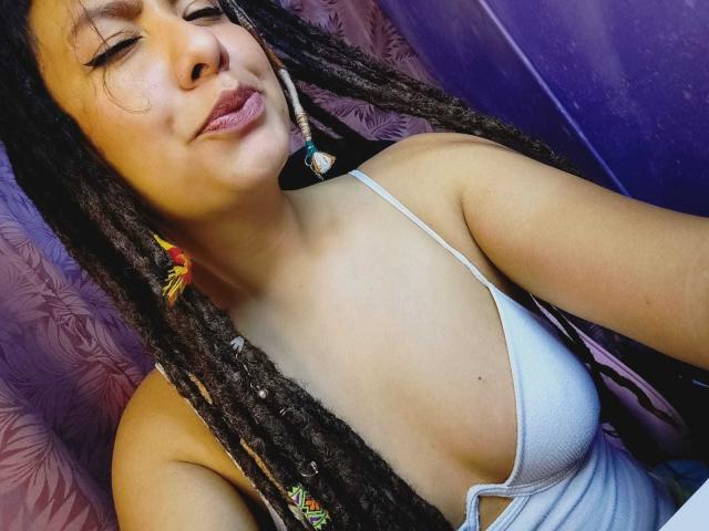 BeckyHuston with dark dreadlocks, pouty lips, and a white tank top, showing her small tits and tan skin. Purple background. Her eyes are closed, smiling slightly.